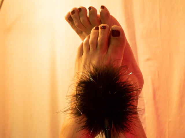RoraimAnaggie's feet with dark red toenails, crossed at the ankles, wearing a fluffy black ankle cuff against a warm, orange-lit background.