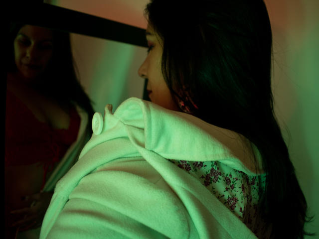 RoraimAnaggie with dark hair, wearing a white hoodie, stares at her reflection in a mirror showing her red bra-covered boobs. Green and red lighting creates a moody effect.