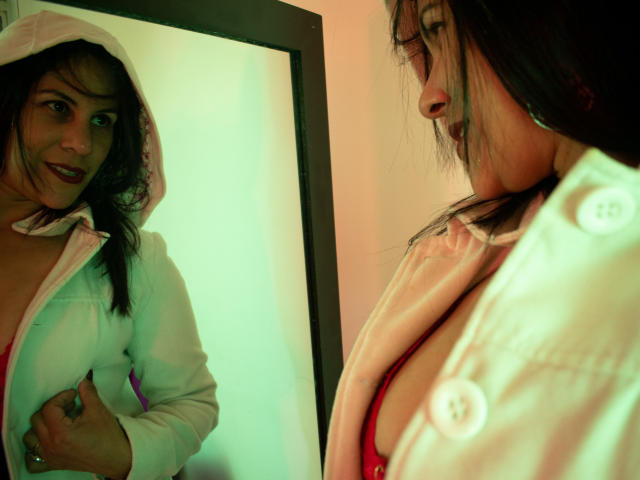 RoraimAnaggie, con cabello oscuro, vestida con una sudadera blanca, un sujetador rojo y botones rosas, se mira en un espejo, mostrando sus pechos y una ligera sonrisa..
