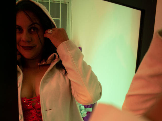 RoraimAnaggie with dark hair, red lace-up corset, and white hoodie, smiling at mirror, hand in hair, showing tits, green and yellow lighting, ring on finger.