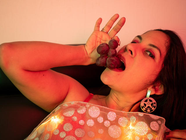 RoraimAnaggie with dark hair, wearing red lingerie and a patterned pillow, eating purple grapes, large hoop earrings, red lighting, tongue out, hand raised to mouth.