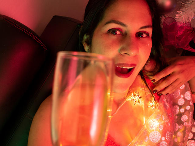 RoraimAnaggie with red lipstick, smiling, holding a glass, wearing a sparkly top, pink and red lighting, dark leather couch, right hand with red nails, relaxed, happy expression.