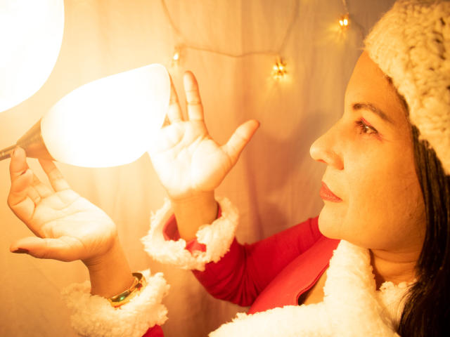 RoraimAnaggie with tan skin, brown eyes, wearing a red and white fluffy Santa outfit, hat, and bracelet, holding hands up to a bright white lamp.