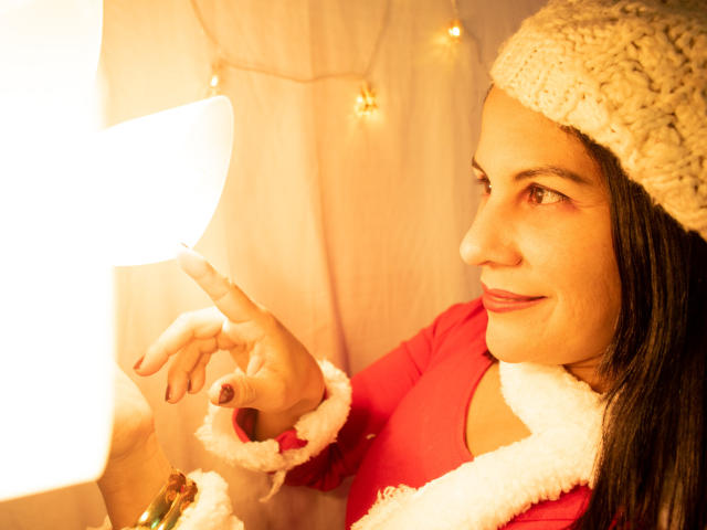 RoraimAnaggie with dark hair, wearing a knitted beige hat and red Santa coat, smiles while pointing at a glowing light. Her tits are partially covered by the coat.