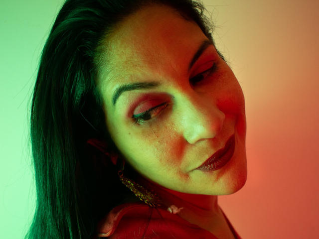 RoraimAnaggie has dark brown eyes with red eyeshadow, freckled face, and dark lipstick, wearing a red shirt, with green and red lighting on her face and dark hair.