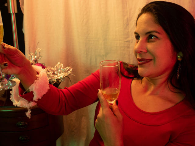 RoraimAnaggie with red shirt, holding glass of wine in right hand, pointing with left at small white ornament on wooden table, smiling, wearing green earrings, dark hair.