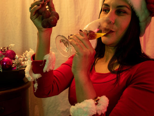 RoraimAnaggie with long dark hair, wearing a red Santa hat and red sweater, sips a sparkling wine, holding grapes in her other hand, looking cute.