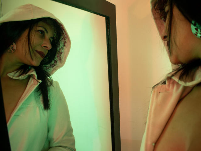 RoraimAnaggie, con cabello oscuro, vestida con una capucha blanca, mirándose en un espejo. Tiene pechos pequeños, una sonrisa encantadora, y se refleja con una iluminación verdosa..