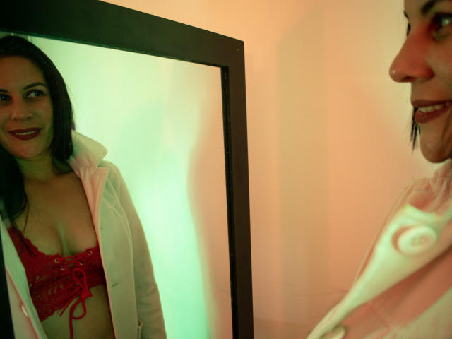 RoraimAnaggie stands in front of a mirror, smiling, wearing a red lace bra and an open white shirt, showing her small tits and dark hair, reflected in the mirror.