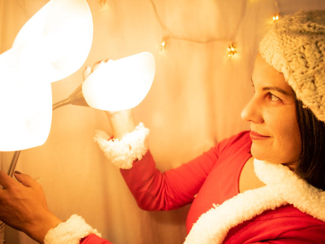 RoraimAnaggie in a red Santa jacket with white fur trim, holding a glowing lamp, wearing a knitted gray hat, smiling, looking to the side, with warm lighting.