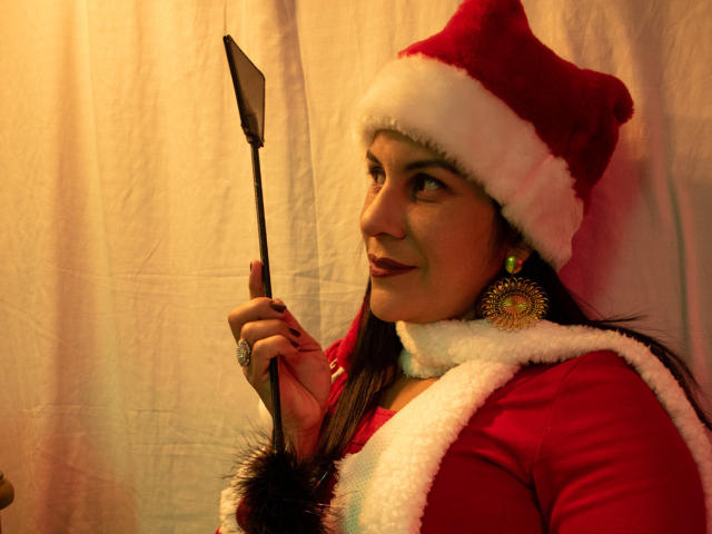 RoraimAnaggie con un gorro de Papá Noel rojo, sosteniendo una pala negra, vestida con un traje de Papá Noel rojo y blanco de piel, sonriendo, con aretes y un anillo, en una foto cálida, con una iluminación ligeramente anaranjada..