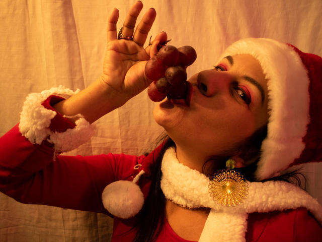 RoraimAnaggie with red makeup, Santa hat, and red dress, sucking on a pair of grapes, wearing a white scarf with a gold brooch, in a warm, yellow light.
