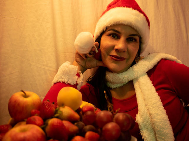 RoraimAnaggie with red clothes and Santa hat, holding a snowball, has a cute smile, wearing a white scarf, and sits with a fruit bowl including apples and grapes.