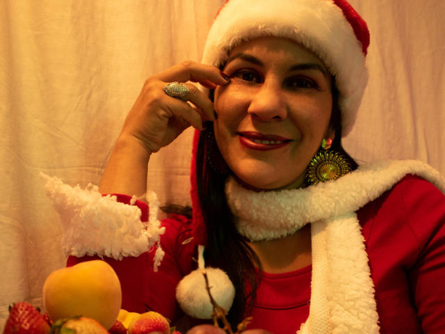 Eine RoraimAnaggie mit brauner Haut, lächelnd, mit einem Weihnachtsmannhut, einem roten Kleid, einem weißen Schal und grünen Ohrringen, die einen glitzernden Ring berührt, neben gelben Äpfeln und roten Dekorationen..
