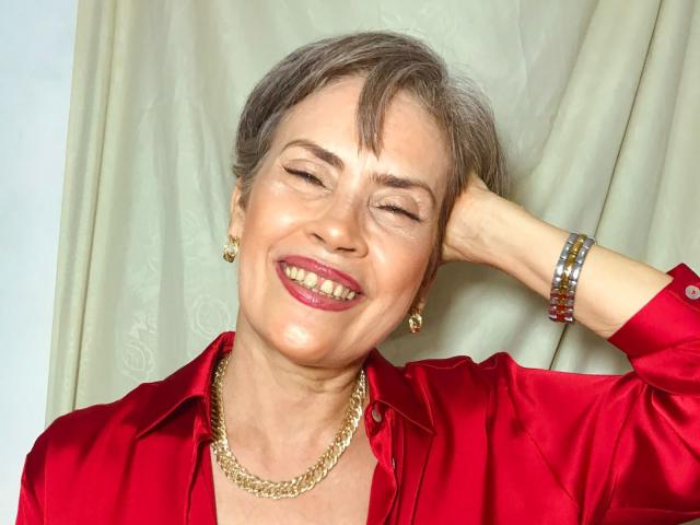 BelleBlondx with gray hair, red lipstick, and a shiny red satin shirt, smiling with eyes closed, hand behind head, gold chain necklace, and red bracelets.