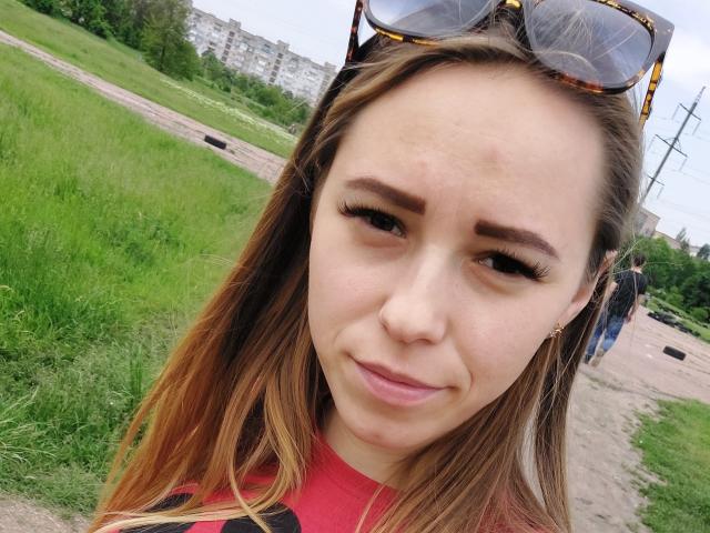 DorrieAnn's face with light brown hair, subtle pout, and thick eyebrows, wearing sunglasses on her head, red shirt, slight redness on her forehead, standing in a grassy area.