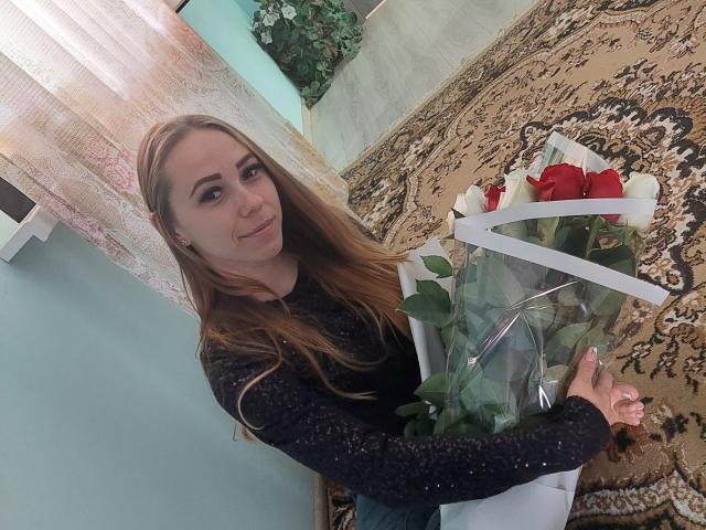 DorrieAnn with long brown hair, wearing a black sparkly top, holding a white basket with red roses and green leaves, standing on a beige patterned rug.