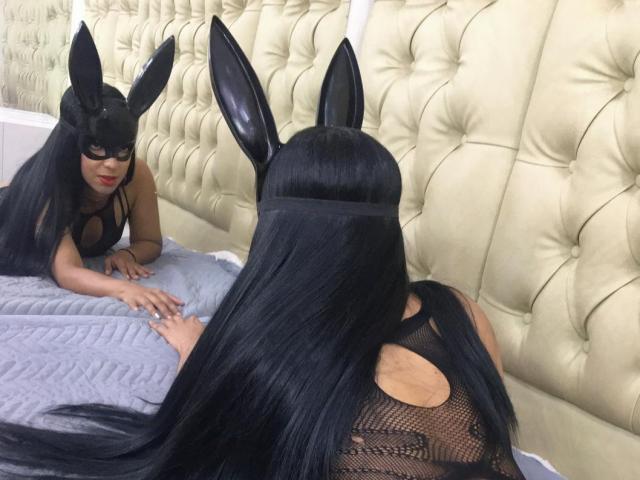 LauraCollingss, com cabelos longos e pretos e orelhas de coelho pretas, usando uma máscara preta e uma blusa de malha, olhando para o seu reflexo em um espelho em uma cama com dossel..