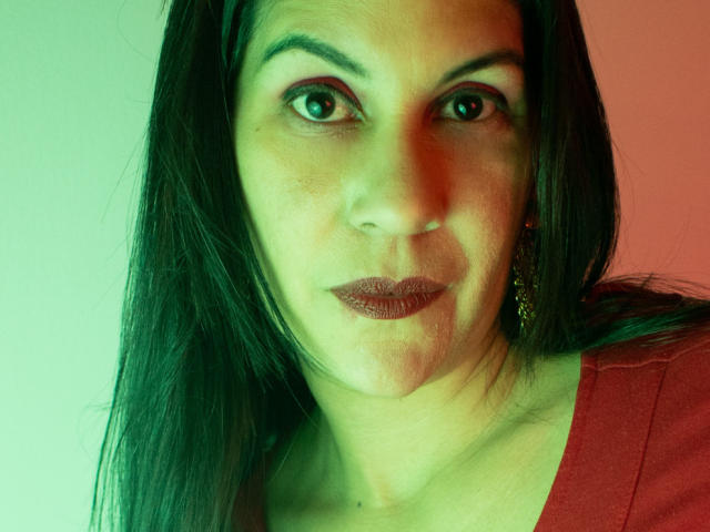 RoraimAnaggie has dark brown eyes, straight black hair, and is wearing dark lipstick with subtle pink eyeshadow. She's wearing a red top and a gold earring.