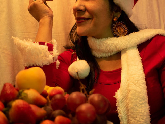 RoraimAnaggie with red dress, white scarf, Santa hat, and gold earrings, holding food near a colorful fruit bowl, smiling, her red lips and dark hair visible.