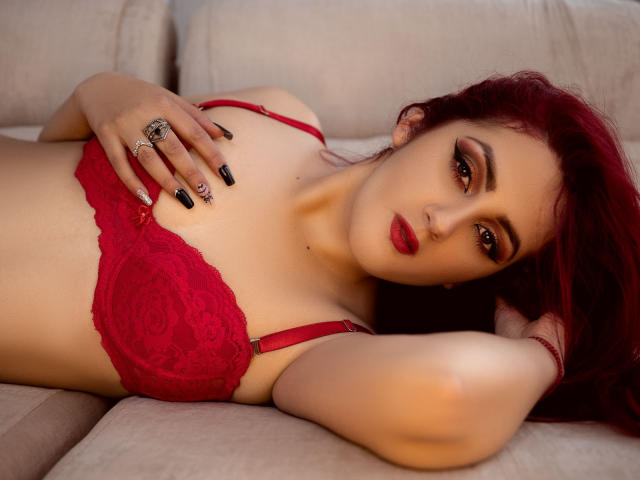 BrunaRousse69 allongée sur un canapé beige, vêtue de lingerie en dentelle rouge, sa main posée sur ses seins, rouge à lèvres rouge, ongles noirs, et cheveux roux, regardant l'appareil photo..