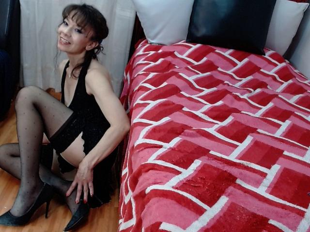 SamaraBellet sitting on wooden floor, wearing black lingerie, sheer polka-dot stockings, black high heels, smiling, next to a red and white patterned bed with black and white pillows.
