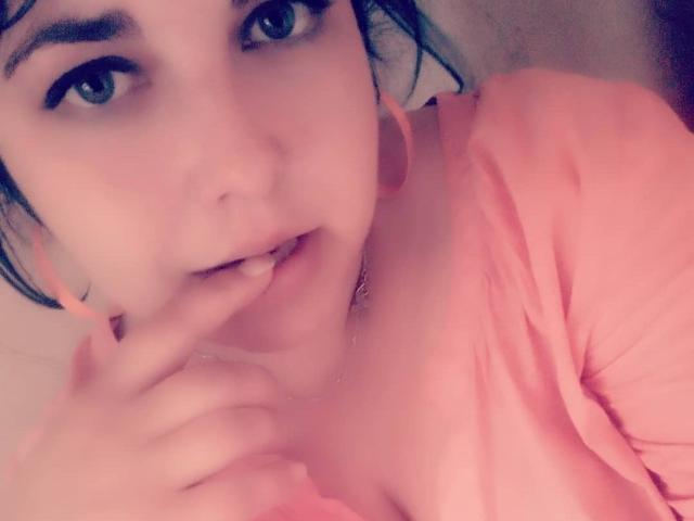 WhitteLotus con ojos azules, camisa rosa y dedo en la boca, con una expresión ligeramente seductora, mostrando un pequeño escote, aros de aro rosas y piel clara. Imagen suave, con un tono rosado, similar a un selfie..