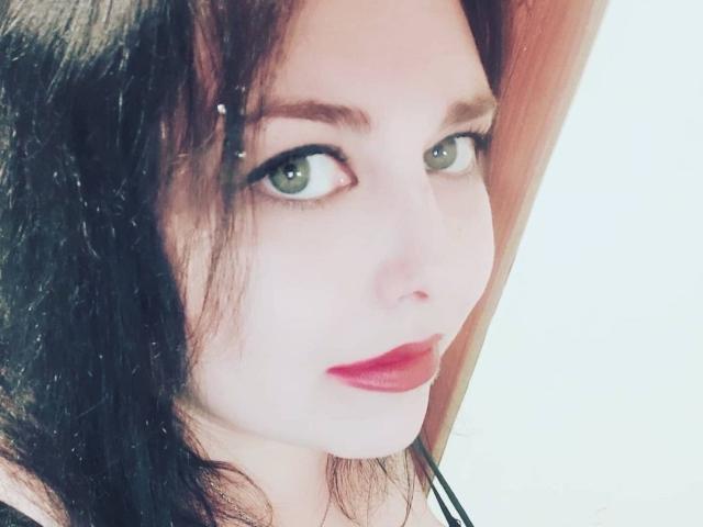 WhitteLotus has pale skin, green eyes, and red lipstick with black eyeliner. She has dark brown hair, a small nose, and a slight smile. Her tits are partially visible.