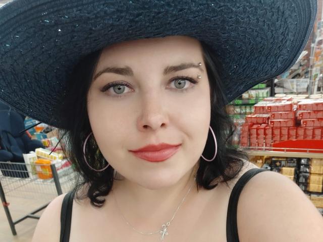 WhitteLotus with pale skin, green eyes, black wavy hair, wearing a black hat, hoop earrings, and red lipstick, has a nose piercing and a cross necklace. Boobs visible.