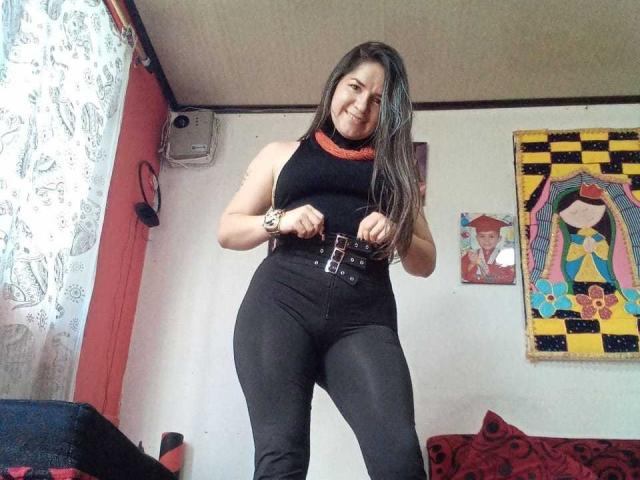 AbbyShiine stands in a room, smiling, wearing a black sleeveless top and high-waisted black pants with a belt, her long brown hair over her shoulder, her tits visible.
