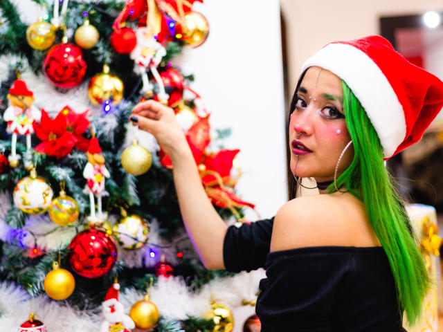 SalomePrince with green hair in a Santa hat, black off-shoulder top, and pink lipstick, reaching for a Christmas tree ornament, featuring red, gold, and white decorations.