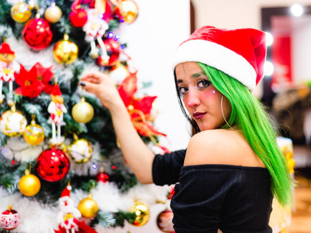 SalomePrince met groen haar, die een kerstmuts draagt en een zwarte top met open schouder, die een versierde kerstboom met rode, gouden en witte versieringen aanraakt, en die omkijkt met een lichte glimlach..