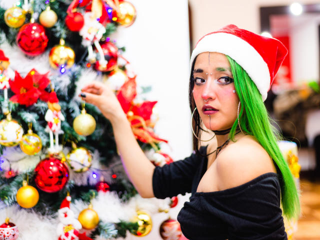 SalomePrince with green hair, Santa hat, black off-shoulder top, large hoop earrings, touching Christmas tree, adorned with red, gold, and white ornaments, in a brightly lit room.