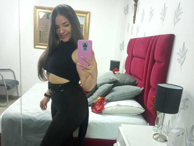 AbbyShiine with long brown hair, smiling, holding pink phone, wearing black crop top, high-waisted black pants, showing her ass, standing in bedroom with red headboard and grey pillows.