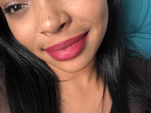Le visage de VickyGreys, avec ses cheveux noirs et lisses, ses yeux marrons, son rouge à lèvres rose, son léger sourire et ses taches de rousseur sur le nez. Un aperçu de cuir bleu turquoise en arrière-plan..