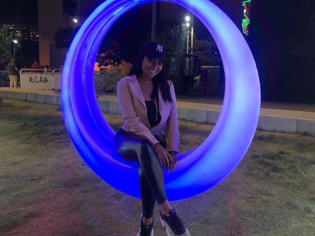 VickyGreys sitting in a glowing blue circular inflatable, wearing a pink jacket, black leather pants, NY cap, and black sneakers, smiling at the camera, night time.