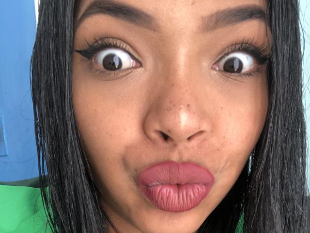 VickyGreys has wide, surprised eyes, dark straight hair, and pink pouty lips. Her brown skin has small freckles and she's wearing green and blue.