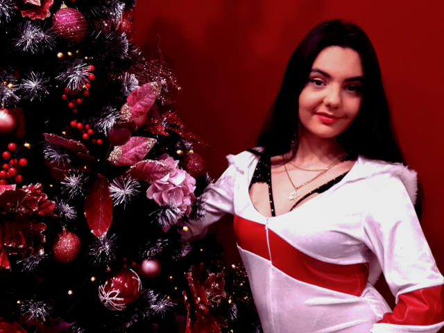 AshlyChloe mit langem, schwarzem Haar, trägt ein weißes und rotes Kleid und steht neben einem geschmückten Weihnachtsbaum mit roten, rosa und silbernen Ornamenten. Ihre Brüste sind sichtbar..