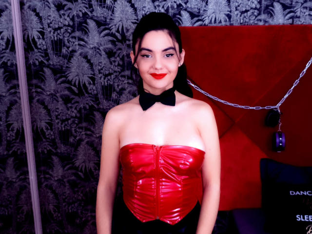 AshlyChloe met een rode, glanzende, mouwloze bustier, een zwarte strik en rode lippenstift, staat voor een rode muur en kijkt met een lichte glimlach naar de camera..