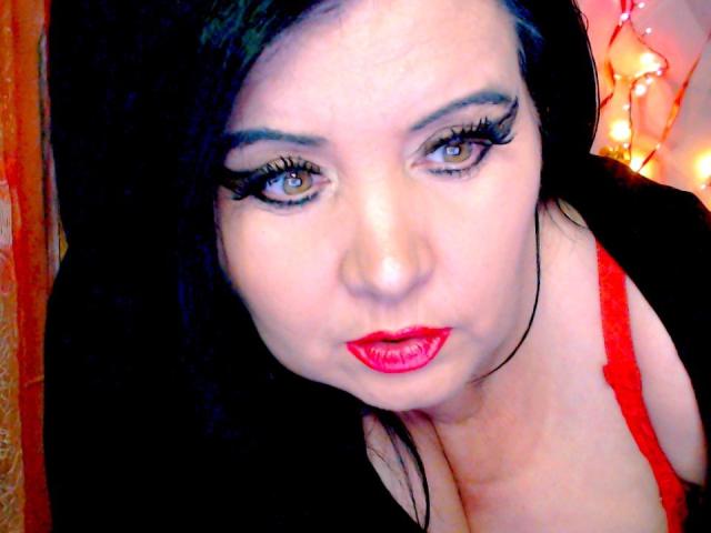 KamilaDream met zwart haar, rode lippenstift en veel eyeliner, kijkt in de camera, draagt een rode bh, toont haar borsten, in een licht schuine selfie..