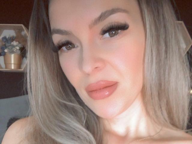 SophieDeee tiene labios carnosos, pestañas largas y cabello rubio, y sus pechos se ven ligeramente en la foto, mirando a la cámara con una sonrisa sutil..