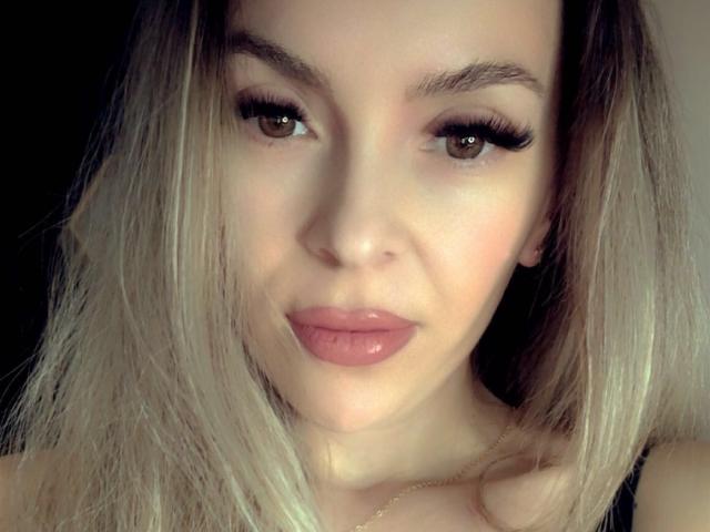 SophieDeee has blonde hair, brown eyes, thick eyebrows, long black eyelashes, and pink glossy lips. Her face is centered with a subtle smile. Her tits are not visible.