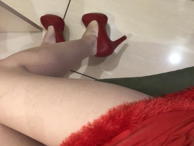 SugarrX's legs in red high heels, wearing a red fluffy dress, sitting on a green chair, pale skin, tiled floor, shadow on the floor, sexy, stylish, seductive.