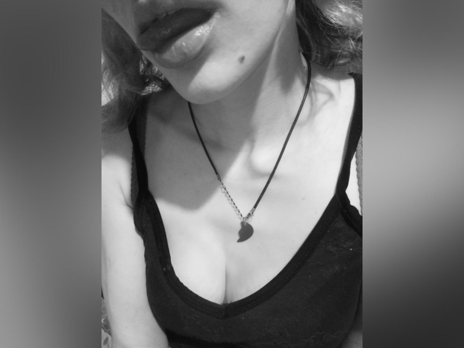 La parte superiore del corpo di Evelyn Kulak, canotta nera, piccola collana a forma di cuore, scollatura, tatuaggio sul braccio destro, capelli biondi ondulati, foto in scala di grigi, labbra leggermente dischiuse, neo visibile sul mento..