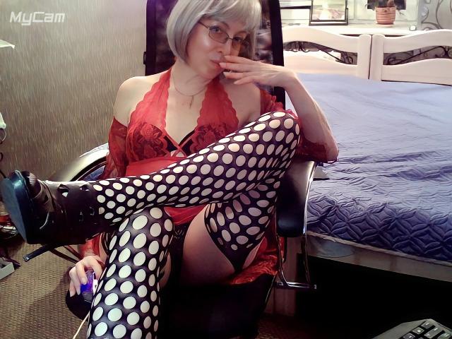 IrunchaArt with gray bob, glasses, red lace lingerie, and polka-dot fishnet stockings, sitting on chair, touching mouth, showing her tits and crossed leg with black boots.