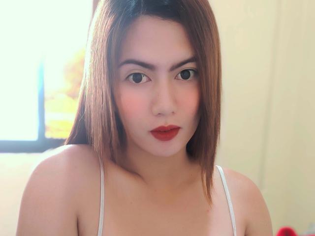 tsHOTLETITIA has straight brown hair, red lipstick, and a white tank top, showing her tits, with a neutral expression, and a small nose piercing.