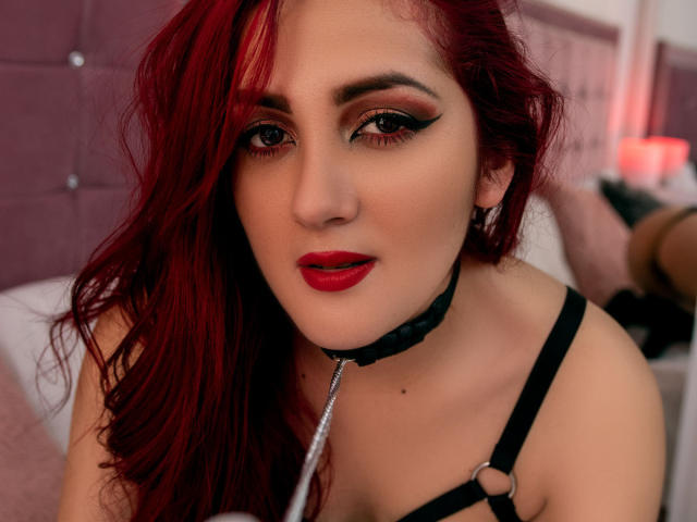 BrunaRousse69 con i capelli rossi, l'eyeliner nero, il rossetto rosso, un'imbracatura nera, un collare in pelle e un guinzaglio argentato, guarda la fotocamera con la bocca leggermente aperta. I suoi seni sono parzialmente visibili..