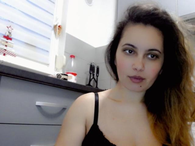 SweetJeniffer with medium tits, wearing a black lace bra, has wavy brown hair, light skin, and a small mole on her chin, in a kitchen with white tiles.