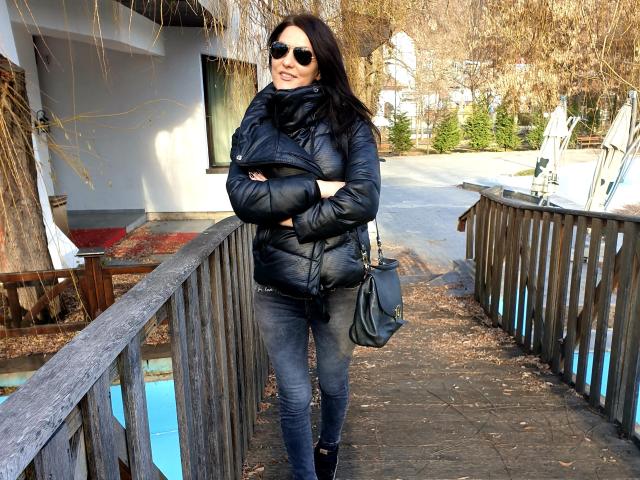 JoliDanielle with black puffy jacket, sunglasses, dark jeans, and black boots, standing on wooden bridge, arms crossed, black handbag, long dark hair, slightly smiling, winter setting.