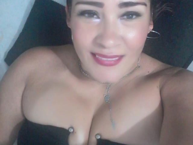 BigSweetGodes with dark hair, hoop earrings, and pink lipstick, smiling at the camera. She has large, shiny tits with diamond studs, wearing a black strapless top.